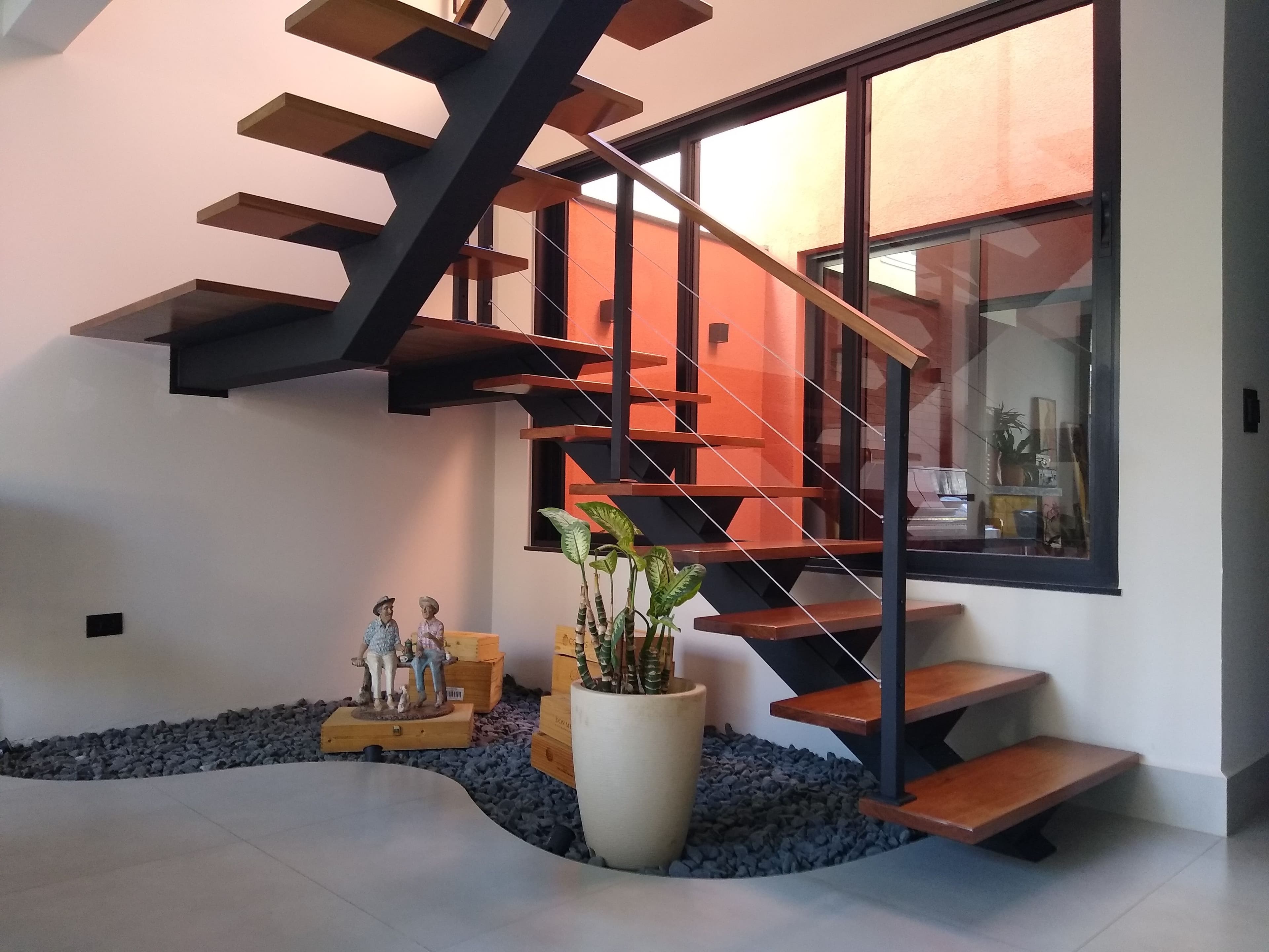 Escada em U com viga central preta, degraus de madeira e cabos de aço em sala de estar com jardim de pedras